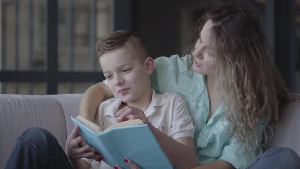 Portrait of a Beautiful Young Mother Reading a Book To Her Beautiful Son Sitting on the Sofa