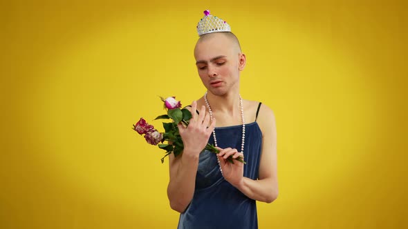 Young Sad Queer Man in Dress and Tiara Holding Bouquet of Withered Flowers Looking at Camera