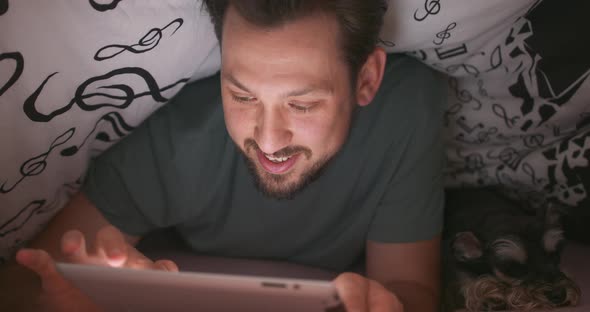 Close Up of Smiling Face of a Young Bearded and Mustashed Man Using Digital Tablet Under Blanket in