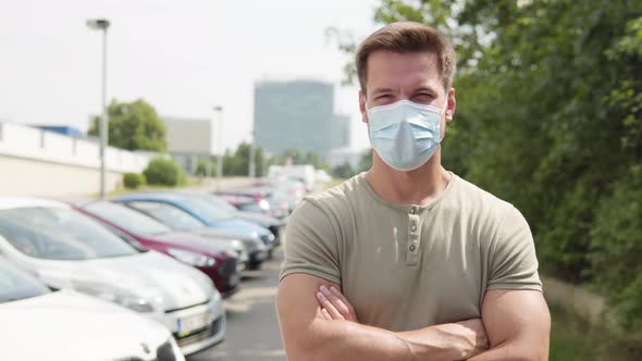 A Young Handsome Man in a Face Mask Looks at the Camera in a Parking Lot in an Urban Area