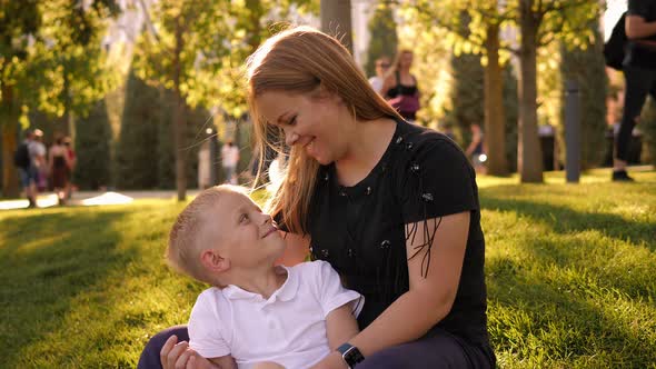 A Mother and Son are Sitting on the Green Grass in the City Park and Hugging