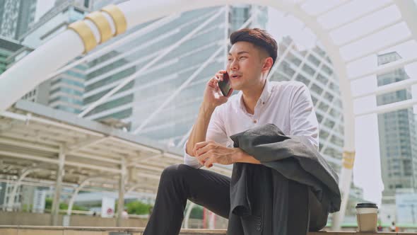 Asian young smart businessman sitting outdoor, talking in telephone with office colleague in city.