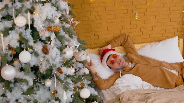 Man in Santa Hat and Sunglasses Lying on Bed Near with Christmas Tree