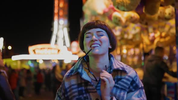 Young Woman Holds Glasses in Teeth at Amusement Park with Bright Lights