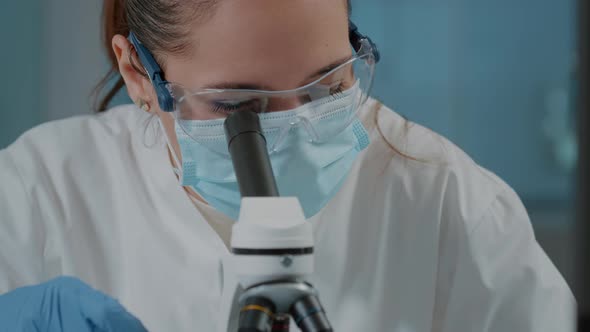 Biologist with Face Mask Using Microscope to Work on Experiment