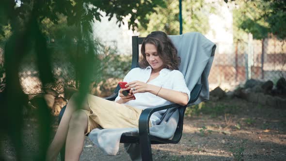 A Young Woman Using Her Smartphone While Sits in an Armchair in the Garden