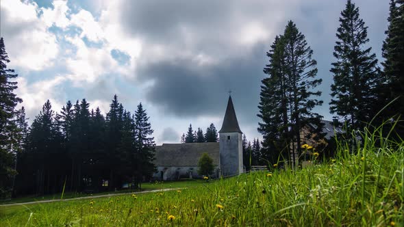 Time lapse, dramatic clouds above alpine village and church and meadow with flowers in front, Trije