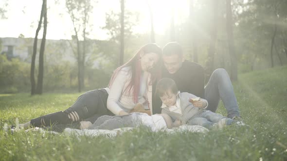 Positive Couple of Young Parents Sitting on Blanket in Sunlight Talking with Cute Little Boy and