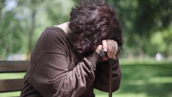 Crying Brunette Senior Woman Sitting with Walking Cane on Bench in Summer Park. Depressed Caucasian