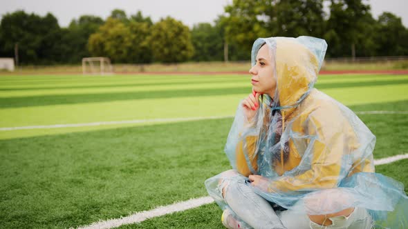  Young woman in raincoat sitting on football field in rainy weather. 