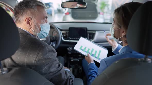 Male Caucasian Dealer in Coronavirus Face Mask Showing Diagrams To Senior Buyer Sitting in New