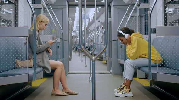 Girls Sitting Opposite Each Other in Train Looking at Phone and Listening Music