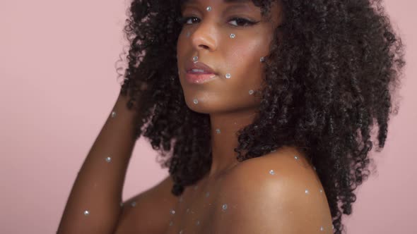 Mixed Race Black Woman with Curly Hair Covered By Crystal Makeup on Pink Background in Studio