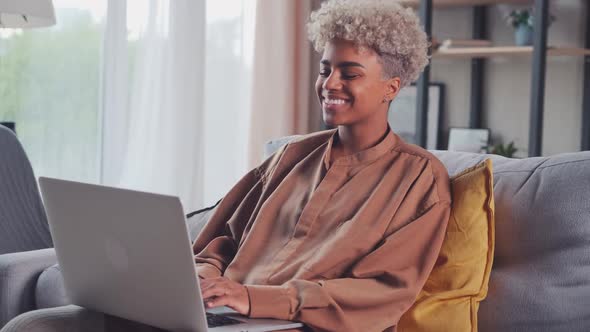 Happy Young African Woman Sitting on Couch Using Laptop Looking at Screen