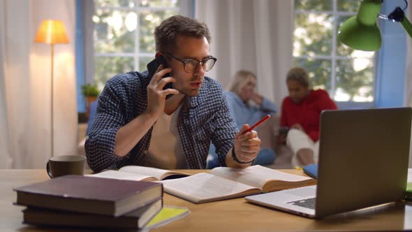 Young Guy Studying on Computer and Talking on Phone with Classmate Preparing for Exam in College