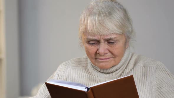 Aged Lady in Wheelchair Holding Book and Looking for Eyeglasses Lying on Table