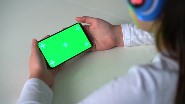 Over Shoulder View of Child in Headphones Holding Smartphone with Green Screen Mockup
