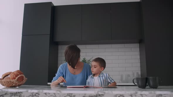 A Young Spanish Mother with Her Son Sitting at the Table Teaches to Read the Child Helping and