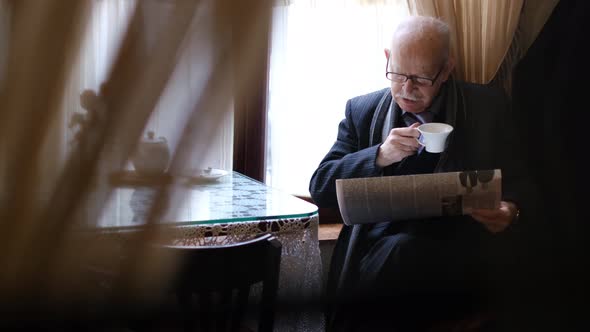 Senior Man Holding a Cup of Coffee Sitting at a Table in a Cafe Reading a Newspaper