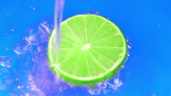 Water pours on green hue juicy lime on blue background slow motion closeup. Lime slice water