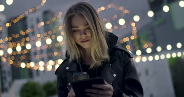 Young Girl in Wireless Headphones Use a Tablet Sitting Outside in a Modern Residential Area, Evening