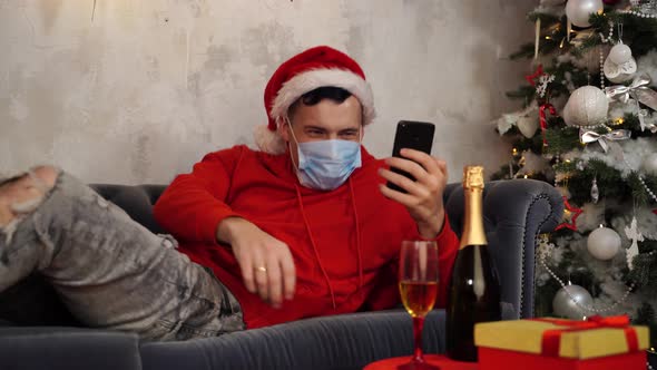 Man in Medical Mask and Santa Hat Conducts Live Broadcast with Friends on Smartphone Lying on Couch
