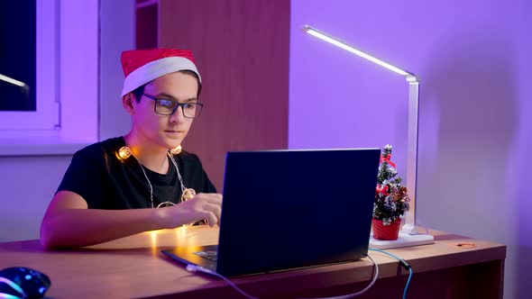 A Young Man in Glasses and a New Year's Hat with a Laptop at the Table. A Student on New Year's Eve