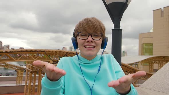 A Young Woman Blogger in Blue Headphones is Broadcasting Outdoor Online and Talking About Events