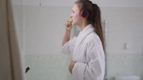 Side View of Young Slim Woman in Bathrobe Brushing Teeth in the Morning in Bathroom at Home
