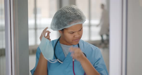 Tired and Exhausted Female African Scrub Nurse Removing Face Mask and Standing Near Window