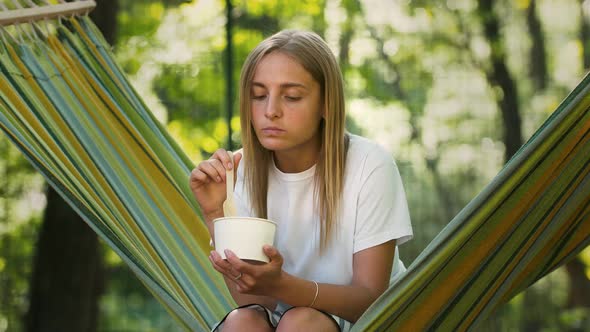 Young Lady is Having a Rest While Sitting in Colorful Hammock and Eating Ice Cream with Fruits