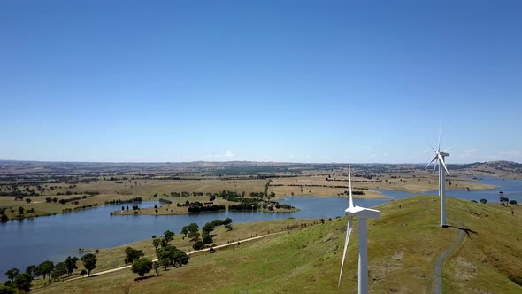 Aerial approach past huge white wind turbines on a grassy green hill beside a meandering river valle alt