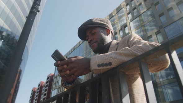 Black Businessman Leaning on Railing and Typing on Smartphone