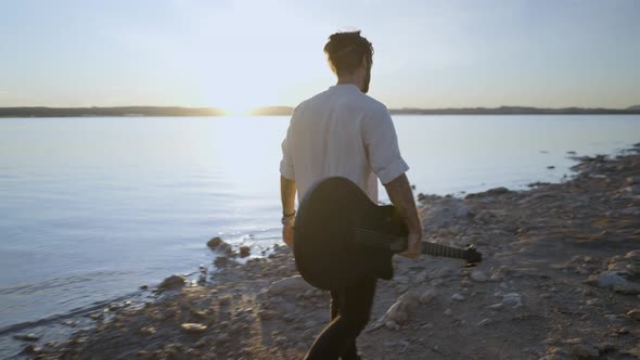 Hipster Man with Guitar Enjoy Sunset at Lake Coast Walking on the Stony Beach Against Sun Slowmotion