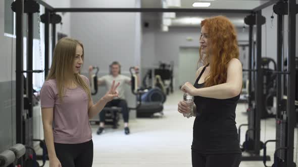 Positive Female Trainer Giving Bottle of Water To the Redhaired Woman in the Gym