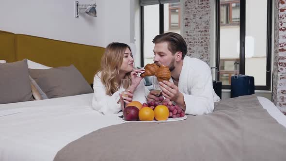 Young Couple in Love Dressed in Bathrobes which Lying on Bed and Girl Feeding Her Boyfreind