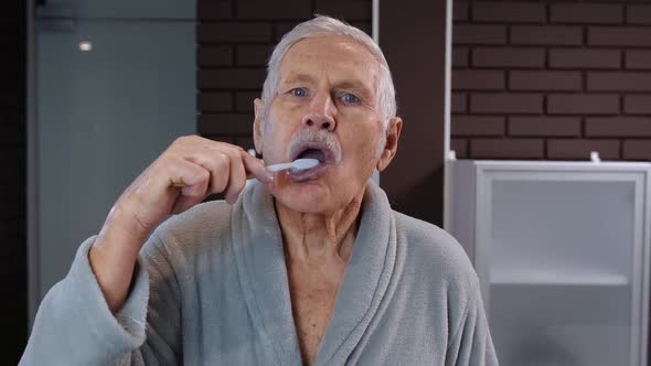 Senior Man Grandfather in Bathrobe Brushing Teeth, Looking Into Mirror. Morning Hygiene at Bathroom