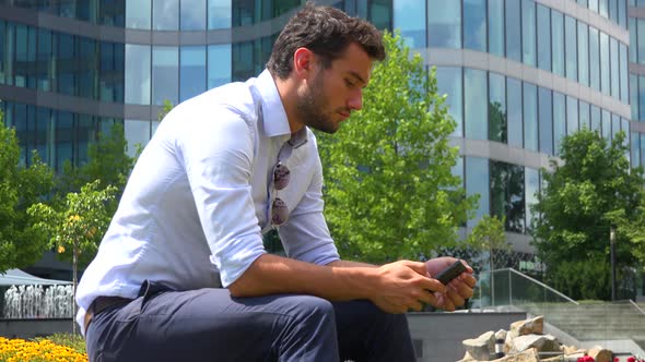 A Young Handsome Businessman Sits in a Park and Works on a Smartphone, an Office Building