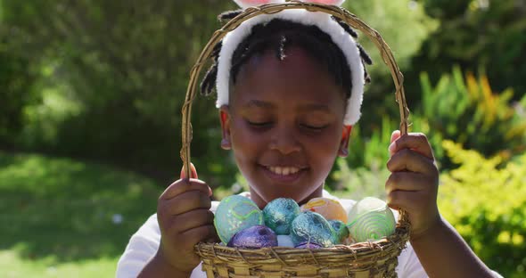 Smiling african american girl wearing easter bunny ears holding basket of easter eggs in garden