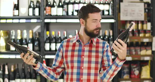 Confused Young Caucasian Man Holding Two Bottles of Wine in Both Hands and Reading Labels. Positive