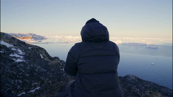 Man In Warm Clothing Kneeling By Camera Tripod On Rocks Of Coastline