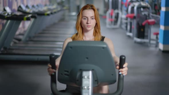 Front View Portrait of Concentrated Fit Caucasian Redhead Woman Riding Exercise Bike in Gym