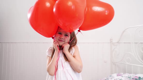 Funny Girl with Red Balloons in Form of Hearts Smiling on Bed By White Wall