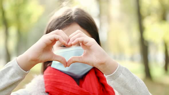 Pretty Girl in Disposable Mask Showing Heart Sign in Autumn Park