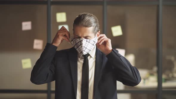 Business Meeting, Young Man in Suit Stands in Conference Room, Man Puts on a Cloth Mask To Protect