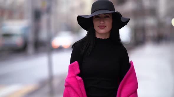 a Brunette in a Black Hat and a Bright Pink Coat Taken Off Shoulders Walks Against the Background of