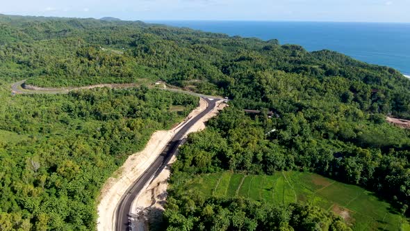 Blue ocean water and winding road under construction in vibrant forest area of Indonesia, aerial vie