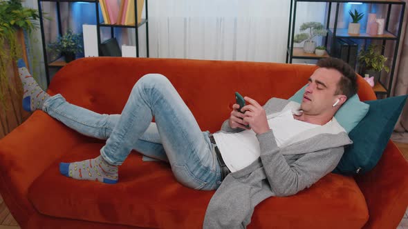 Happy Relaxed Man in Wireless Earphones Dancing Listening Favorite Music Lying on Couch at Home