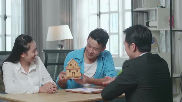 Asian Couple With A House Purchase Contract Paper Receives The House Model From A Real Estate Agent