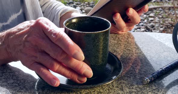 Close up on an elderly woman reading a book and drinking a cup of herbal tea in the morning sun.